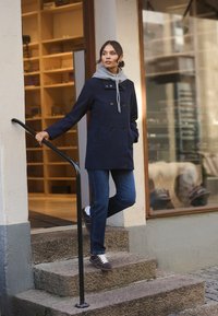 Marineblauer doppelt-knöpfbarer Mantel mit Kapuze, kombiniert mit blauen Jeans und grauen Turnschuhen, stehend auf Steinstufen vor einem Geschäft.
