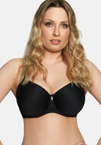 Donna bionda con capelli mossi che indossa un reggiseno imbottito nero con ferretto, in posa su uno sfondo chiaro e uniforme.
