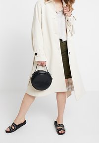 Manteau crème, chemise à carreaux blanche, shorts vert olive, sac à main noir circulaire, sandales noires à clous ; design minimaliste, textures variées.