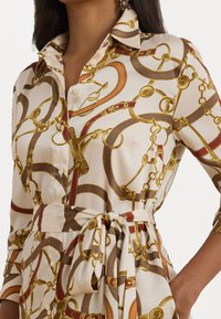 Abito camicia beige chiaro con stampa a catena e cintura in oro, rosso e marrone. Con colletto, vita annodata e chiusura a bottoni.