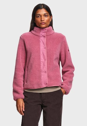 Frau trägt eine pinke Fleecejacke mit Druckknöpfen und steht mit einer Hand in der Tasche vor einem einfarbigen hellen Hintergrund.