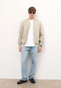 Heled beez värvi bomber-jakk, valge T-särk, heledad sinised teksad ja mustad tossud. Modell kannab tumedaid prille ning seisab ühtlase tausta ees.