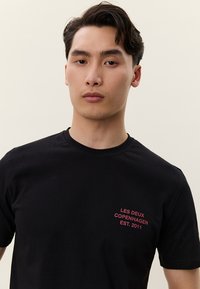 Mladý muž s krátkými tmavými vlasy, oblečený v černém tričku s růžským textem "LES DEUX COPENHAGEN EST. 2011" na hrudi, na neutrálním pozadí.