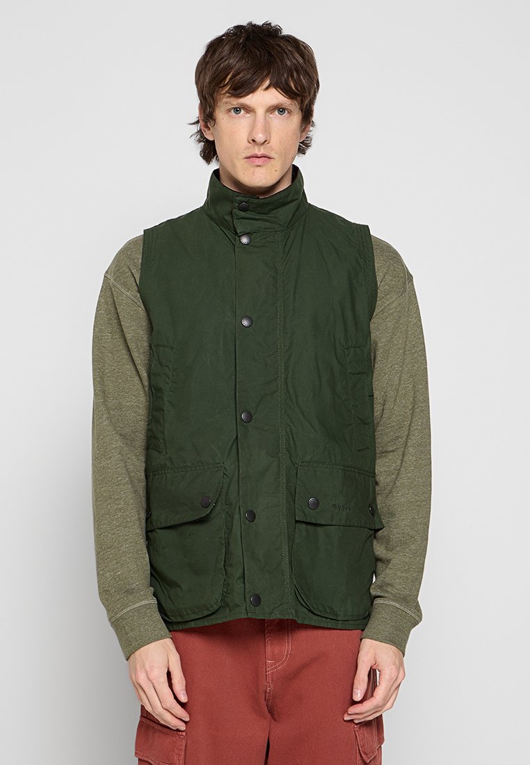 Junger Mann mit kurzen braunen Haaren, der eine dunkelgrüne, knöpfbare Weste über einem olivfarbenen Langarmhemd und rotbraunen Hosen trägt, vor einem weißen Hintergrund stehend.