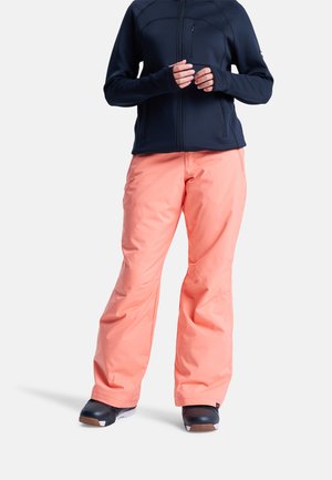 Pantalones de nieve rosas de corte holgado, combinados con una chaqueta azul oscuro con cremallera y agujeros para los pulgares. El conjunto se completa con botas de nieve negras.
