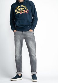 Granatowa bluza z kapturem z grafiką na przodzie w kolorze żółtym i różowym, w połączeniu z jasnoszarymi jeansami i granatowymi butami wsuwanymi z białą podeszwą.