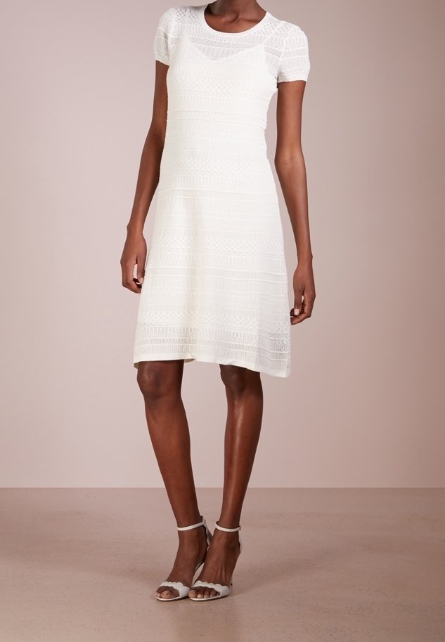 Femme portant une robe blanche en dentelle longueur genou avec manches courtes et des sandales blanches à talons avec bride à la cheville, debout devant un fond uni.