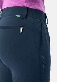 Gros plan sur un pantalon d'extérieur bleu marine avec une poche arrière zippée, une ceinture texturée noire et le logo « Vaude », porté avec un haut violet.