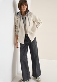 Femme appuyée contre un mur, portant un blazer crème à double boutonnage avec une capuche grise, une chemise blanche, un jean noir large et des baskets avec des accents verts.