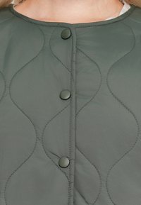 Gros plan sur une veste grise matelassée avec des boutons ronds et un motif de couture ondulé sur le tissu, portée sur un haut de couleur claire.