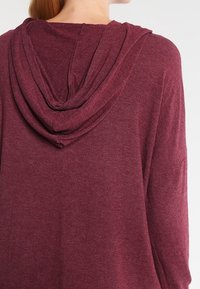 Maroon hooded top made of soft fabric with a relaxed fit. The back features a draped hood detail and long sleeves, offering casual comfort.