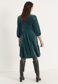 Teal fluwelen jurk met driekwartmouwen, een gelaagde rok en een geraffineerde taille, gecombineerd met kniehoge zwarte laarzen en doorschijnende tights.