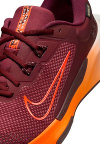 Gros plan sur une sneaker bordeaux avec une semelle orange, un tissu texturé, un swoosh souligné d'orange et des lacets bordeaux.