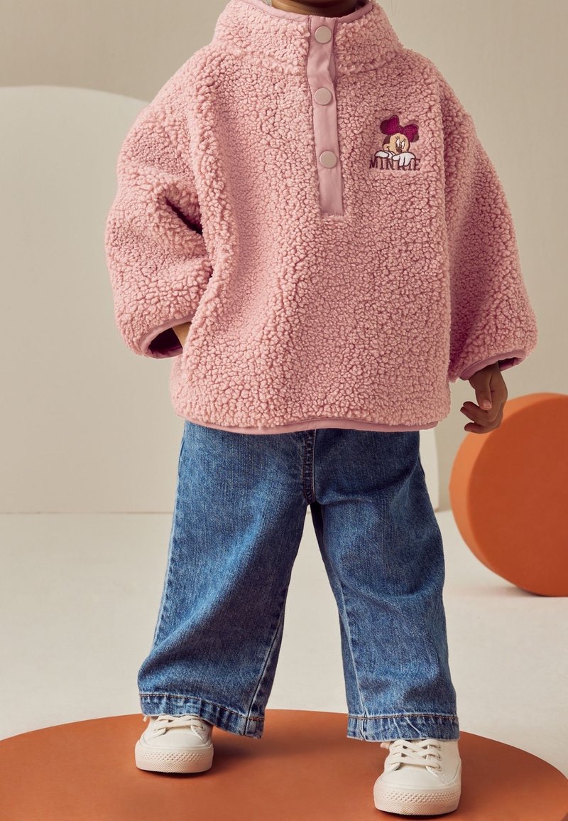 Enfant portant un pull en polaire rose oversize avec le logo Minnie Mouse, un jean large bleu et des baskets blanches, debout sur un cercle orange.