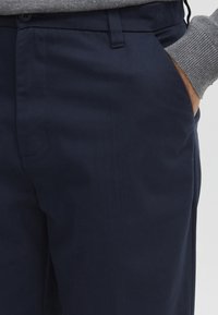 Mand iført navy blå bukser og grå sweater med højre hånd i bukselommen, beskåret ved talje og lår.