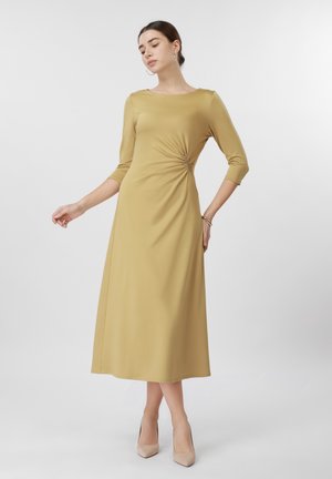 Vrouw draagt een mosterdgele midi-jurk met driekwartmouwen en geplooide details bij de taille, gecombineerd met beige hakken tegen een effen achtergrond.