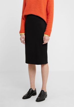 Person wearing a knee-length black skirt, orange sweater with yellow cuffs, and black lace-up shoes standing against a white background.