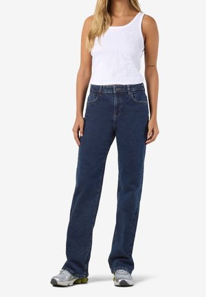 Kvinde iført en hvid ærmeløs ribstrikket top, mørkeblå jeans med lige ben og sølvfarvede sneakers, stående foran en ensfarvet baggrund.