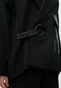 Čierny vonkajší odev z textúrovaného vlneného zmesi, so širokým golierom, voľným strihom a koženým vreckom s kruhovým kovovým doplnkom.