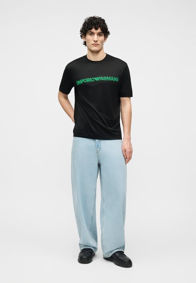 Νεαρός με σγουρά μαλλιά που φοράει μαύρο T-shirt Emporio Armani και ανοιχτόχρωμα φαρδιά τζιν, στέκεται με το ένα χέρι πίσω από την πλάτη του.