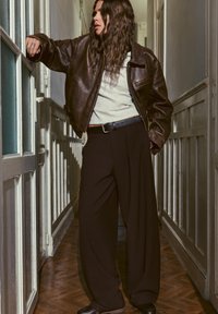 Femme aux cheveux ondulés portant une veste en cuir marron, un haut blanc et un pantalon large noir, appuyée contre une porte dans un couloir étroit.