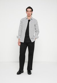 Fekete-fehér mintás, gombos hosszú ujjú ing, fekete póló fölött viselve; fekete nadrággal és cipővel párosítva.