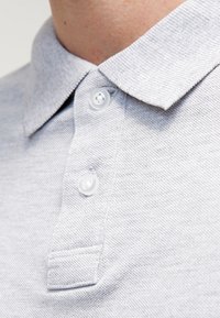 Polo gris en tissu texturé, col côtelé et trois boutons blancs. Vue en gros plan mettant en évidence les coutures et les détails des boutons.