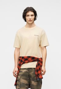 Νεαρός άνδρας με μπεζ T-shirt McLaren, παντελόνι καμουφλάζ cargo και κόκκινο-μαύρο καρό πουκάμισο δεμένο γύρω από τη μέση του, στέκεται μπροστά σε ένα απλό φόντο.