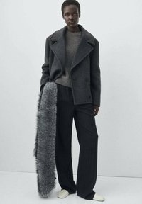 Cappotto in lana grigio con ampi revers, indossato sopra un maglione grigio chiaro. Pantaloni a righe grigie a gamba larga e una lunga sciarpa di pelliccia sintetica grigia.