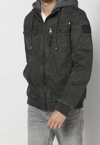 Homme portant une veste zippée vert foncé avec capuche et plusieurs poches frontales, associée à un jean gris clair.