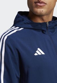 Marineblauwe hoodie gemaakt van gladde stof. Heeft witte drie-strepen accenten op de schouders en een geborduurd wit logo op de borst.