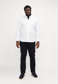 Camicia bianca a maniche lunghe con bottoni abbinata a pantaloni neri e sneakers nere, caratterizzata da un colletto classico e design con bottoni frontali.