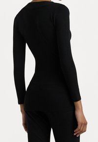 Mujer vistiendo una blusa negra ajustada de manga larga y pantalones negros, vista desde atrás contra un fondo blanco liso.