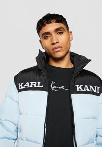 Polstret jakke med en lyseblå underdel og svart overdel. Har logoene "KARL" og "KANI" på brystet. Sort glidelås.
