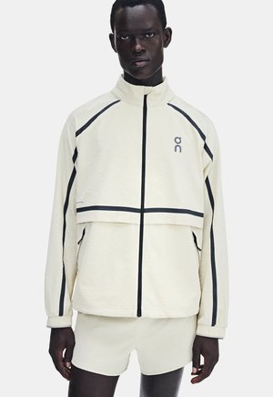 Homme portant une veste polaire blanche zippée et un short assorti avec des bandes noires sur les manches et la poitrine.