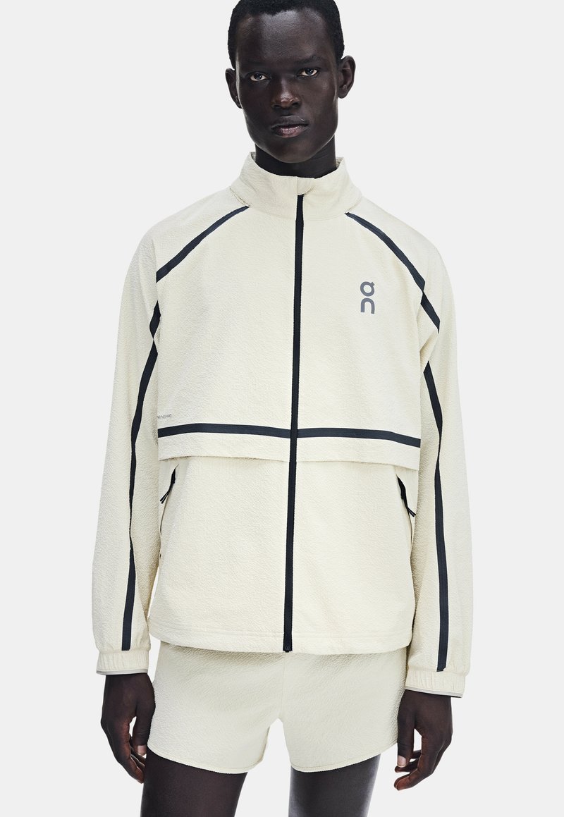 Homme portant une veste polaire blanche zippée et un short assorti avec des bandes noires sur les manches et la poitrine.