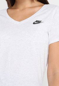 Camiseta de algodón blanca con un diseño de cuello en V. Presenta un pequeño logo negro de Nike en el lado izquierdo del pecho. Manga corta y corte recto. Textura suave.