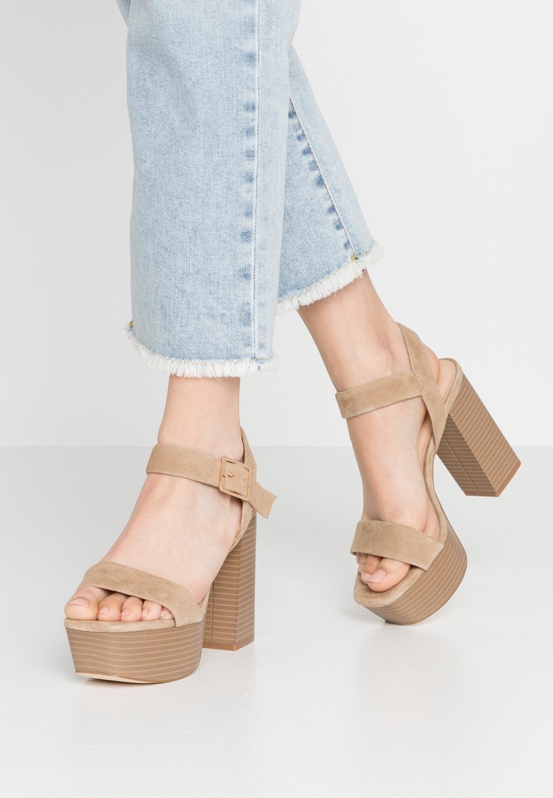 Pieds portant des sandales à plateforme en daim beige avec talons blocs, associées à un jean bleu clair coupé à l'ourlet effiloché, sur fond blanc.