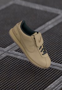 Adidași Nike bej, cu un part superior din piele netedă, căptușeală interioară verde și șireturi negre. Au o limbă căptușită și o talpă din cauciuc texturat.