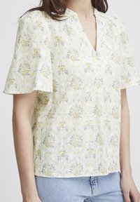 Blus med blommönster i ljusgult med korta, svävande ärmar. Har en V-ringning och subtila veck. Mjuk, texturerad tyg.