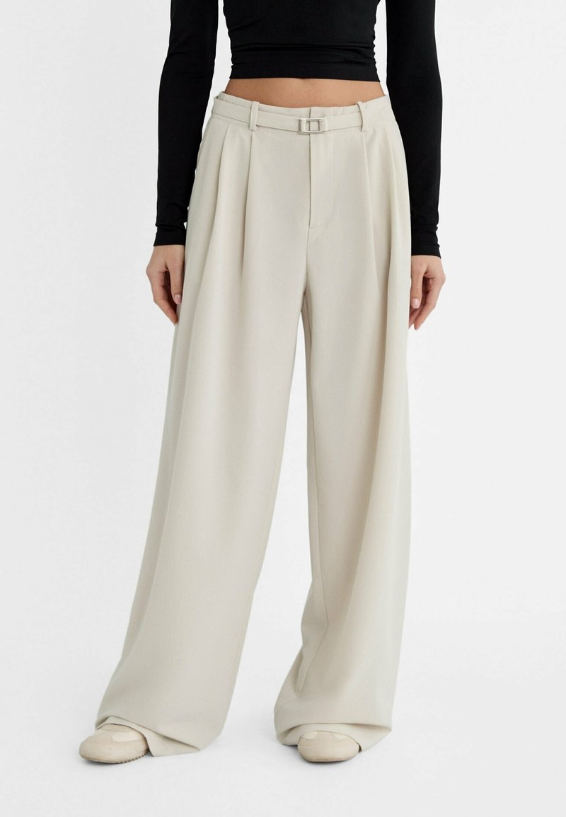 Pantalons larges beige avec des plis, une taille ajustée et un détail de boucle en argent. Tissu lisse avec un léger éclat.
