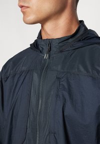 Veste légère navy avec capuche, dotée d'une fermeture éclair frontale intégrale, de sections en tissu texturé et d'un design minimaliste.