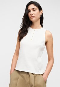 Άσπρο αμάνικο τοπ με στρογγυλή λαιμόκοψη, διακοσμημένο με πέντε χρυσά κουμπιά στο μπροστινό μέρος. Κατασκευασμένο από απαλό ύφασμα, με μικρή λεπτομέρεια λογοτύπου στο τελείωμα.