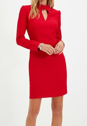 Kobieta w dopasowanej, czerwonej sukience z długim rękawem, z niewielkim wycięciem z przodu oraz srebrną bransoletką na lewej ręce.