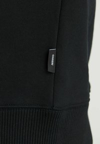 Sweat-shirt noir avec poignets côtelés, tissu à texture lisse et une petite étiquette affichant "CHASIN'" sur le côté gauche.