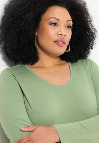 Halenka s dlouhým rukávem v jemně zelené barvě, s véčkovým výstřihem a hladkou texturou. Modelka s kudrnatými černými vlasy, doplněná zlatými náušnicemi.