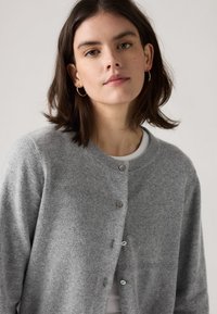 Junge Frau mit schulterlangem dunklem Haar, die goldene Creolen trägt und eine graue Strickjacke mit Knöpfen über einem weißen Oberteil vor einem schlichten Hintergrund trägt.