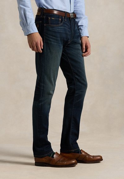 Bărbat purtând blugi slim fit albastru închis, cămașă bleu cu nasturi, curea maro din piele, șosete închise la culoare și mocasini maro din piele, stând în picioare.