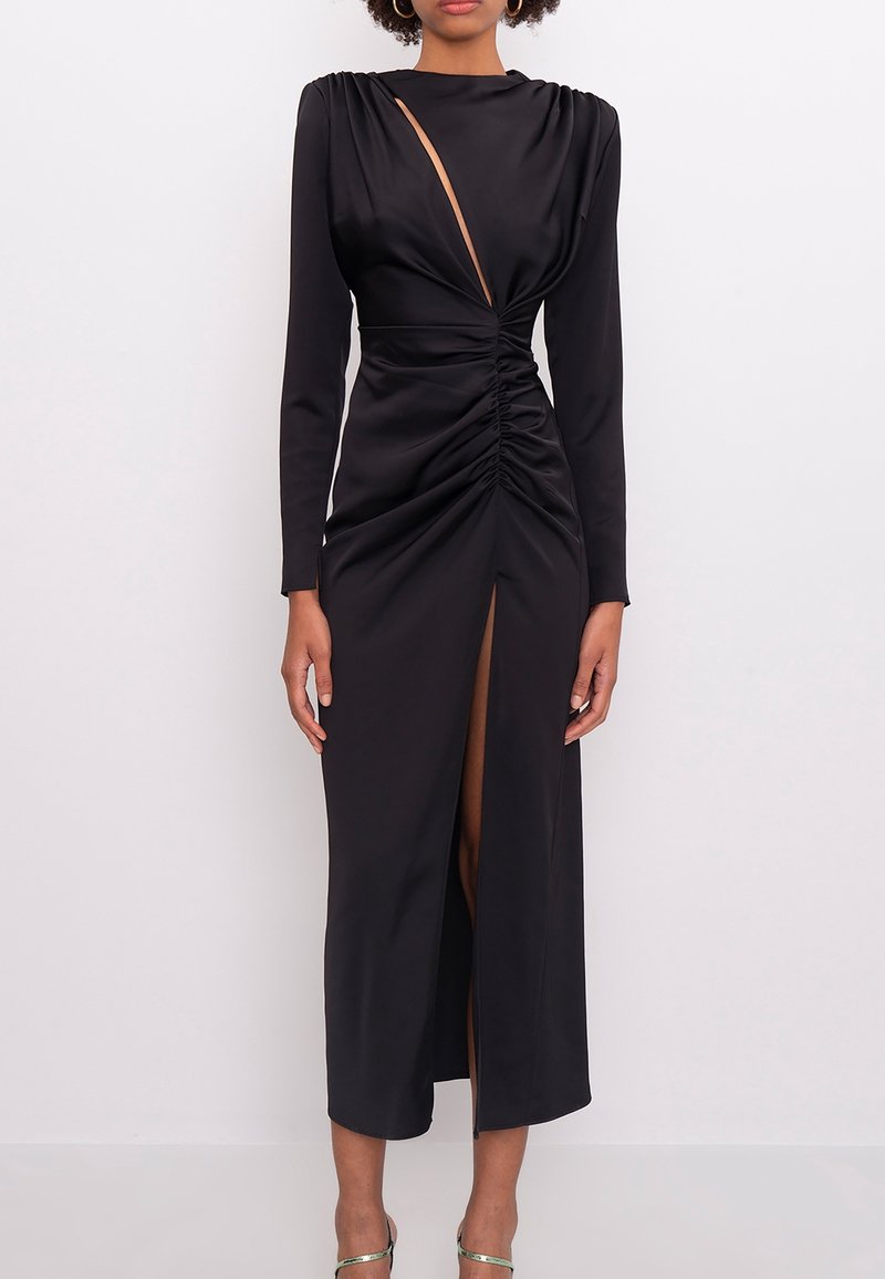 Robe midi noire à manches longues avec taille froncée, fente avant et découpe asymétrique près de l'encolure portée par une femme debout.