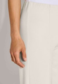 De linkerhand van een vrouw ligt ontspannen naast lichte beige broek met zichtbare verticale naden aan de zijkant.